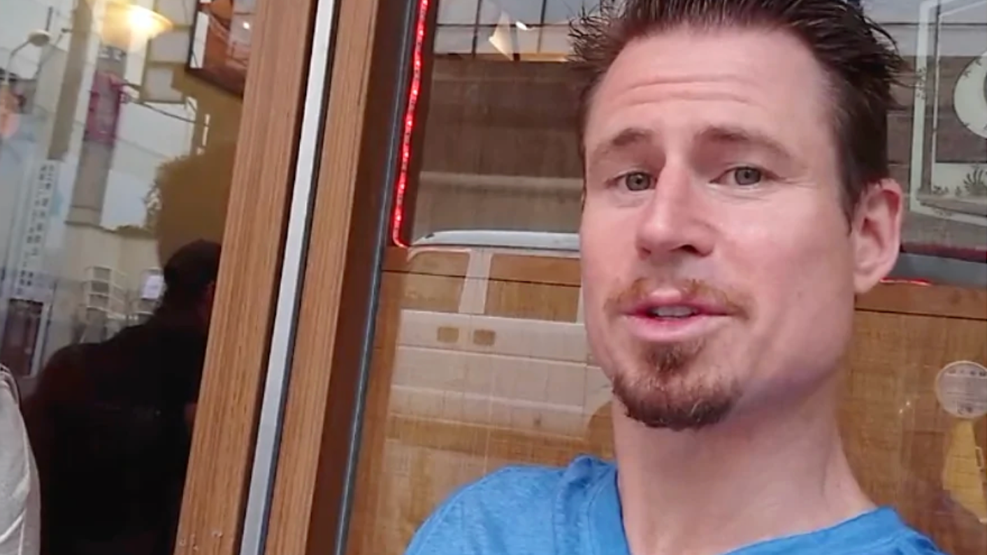 Marcus Talks about Crack The Female Code | #1 Dating Tips and Relationship Coaching Book for Men and How Powerful it really is! | Karl is wearing a Blue T Shirt Sitting outside a Cafe leaning up against the wall.